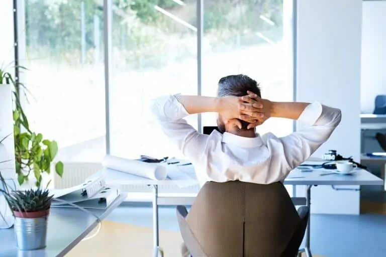 Handsome young businessman in his office, sitting at the desk, stretching arms. Rear view.